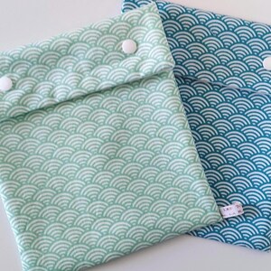 May include: Two fabric pouches with a repeating white wave pattern. One pouch is light mint green and the other is teal blue. Each pouch features a fold-over flap secured by two white plastic snap buttons and a small white fabric tag on the side.