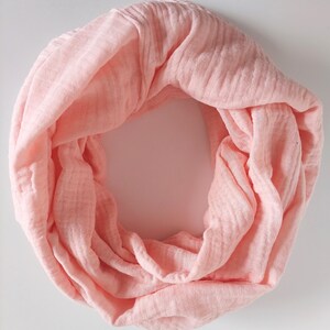 May include: A pale pink infinity scarf made from a textured material. The scarf is in a continuous loop, with a draped and flowing design. The fabric has a subtle, crinkled appearance, giving a relaxed and comfortable feel.
