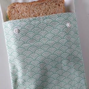 May include: A rectangular fabric sandwich bag featuring a light green and white repeating fan-shaped pattern. The pouch is secured with two white plastic snap buttons at the top and contains a single slice of whole wheat bread inside the bag.