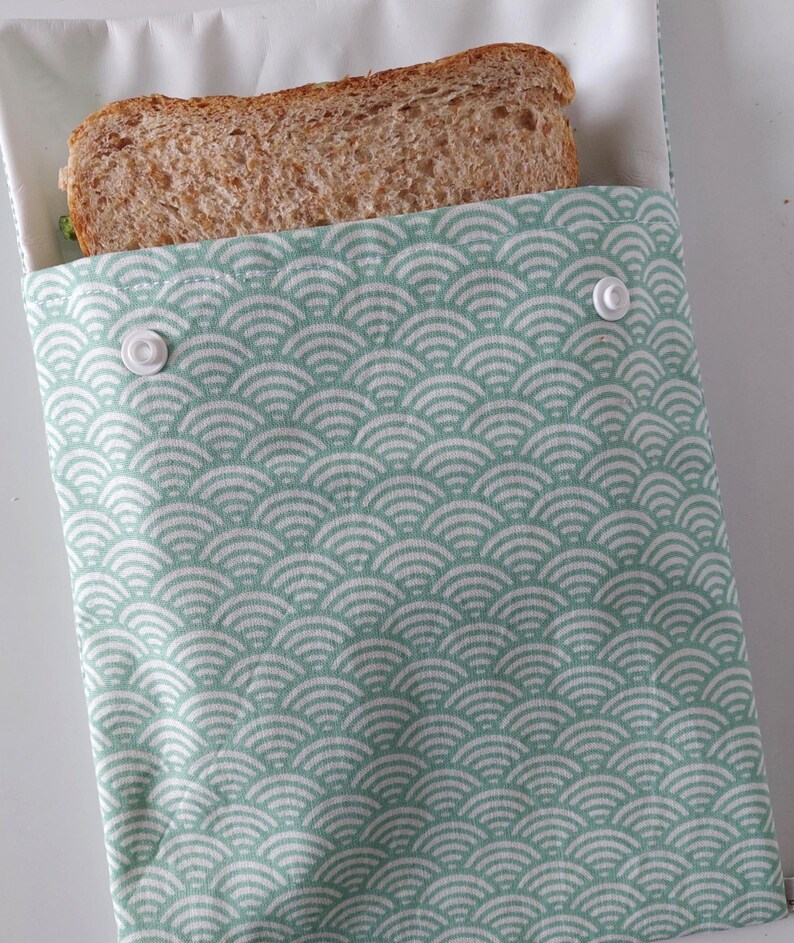 May include: A rectangular fabric sandwich bag featuring a light green and white repeating fan-shaped pattern. The pouch is secured with two white plastic snap buttons at the top and contains a single slice of whole wheat bread inside the bag.