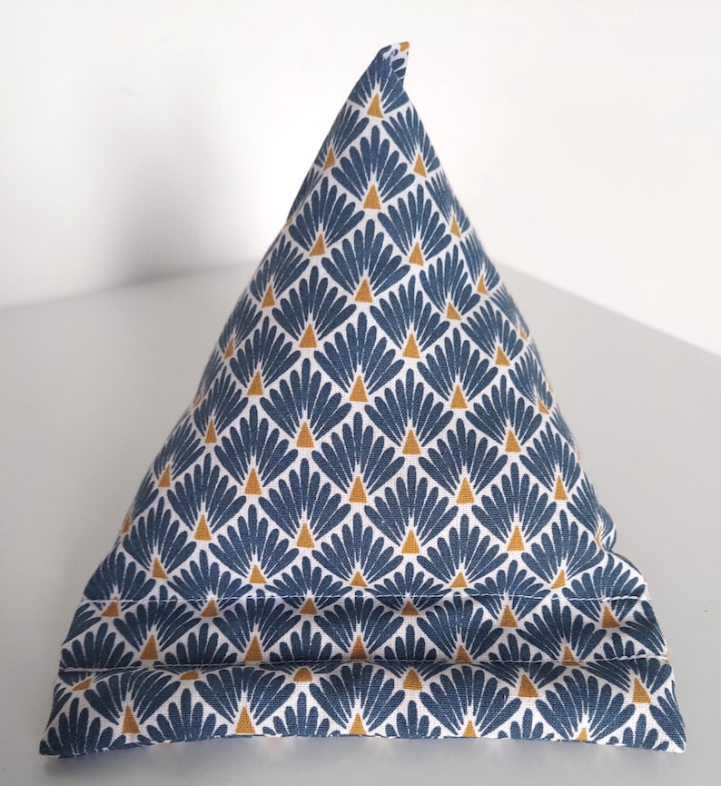 May include: A pyramid-shaped fabric tablet stand featuring a repeating pattern of blue fan-like shapes and small yellow triangles on a white background. The base includes two horizontal stitched ridges designed to hold a device in place.