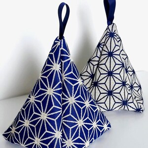 Op de afbeelding: Twee piramidevormige stoffen gewichten met een geometrisch sterpatroon. Eén gewicht heeft een blauwe achtergrond met witte lijnen, en de andere heeft een witte achtergrond met blauwe lijnen. Elk stuk heeft een blauw lintlusje aan de bovenkant.