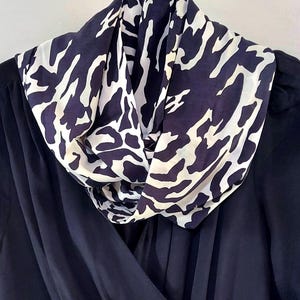 May include: A navy blue scarf with a white animal print pattern is draped over a dark blue garment. The scarf is made of a lightweight fabric and is styled in a cowl neck design. The garment appears to be a dress or top.