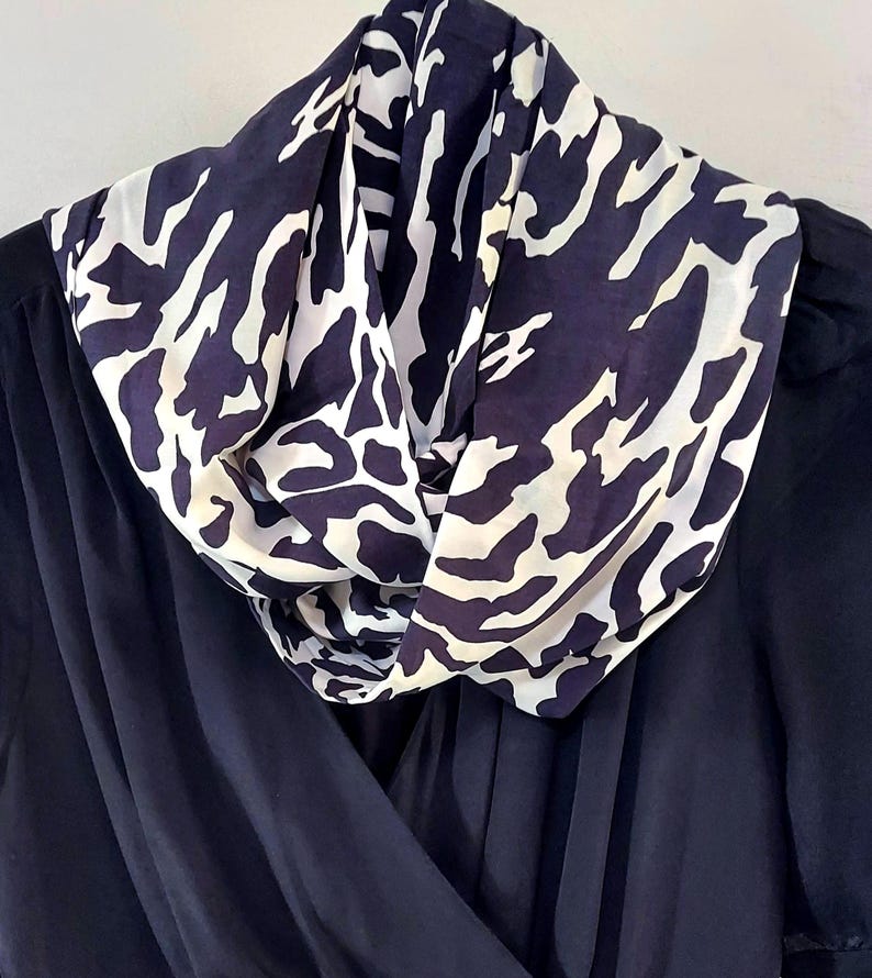 May include: A navy blue scarf with a white animal print pattern is draped over a dark blue garment. The scarf is made of a lightweight fabric and is styled in a cowl neck design. The garment appears to be a dress or top.
