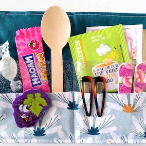 May include: A teal fabric organiser with a floral print pocket, containing a wooden spoon, pink candy, tea packets, hair clips, and nail files. The candy wrapper reads "MAOAM Stripes". A grape-shaped sticker is also visible.