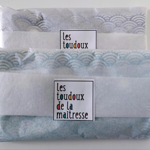 May include: Two rectangular items wrapped in white tissue paper with a blue and white wave pattern visible through the paper. Each package features a white label with the text "les toudoux" or "les toudoux de la maitresse" in black and colorful letters.