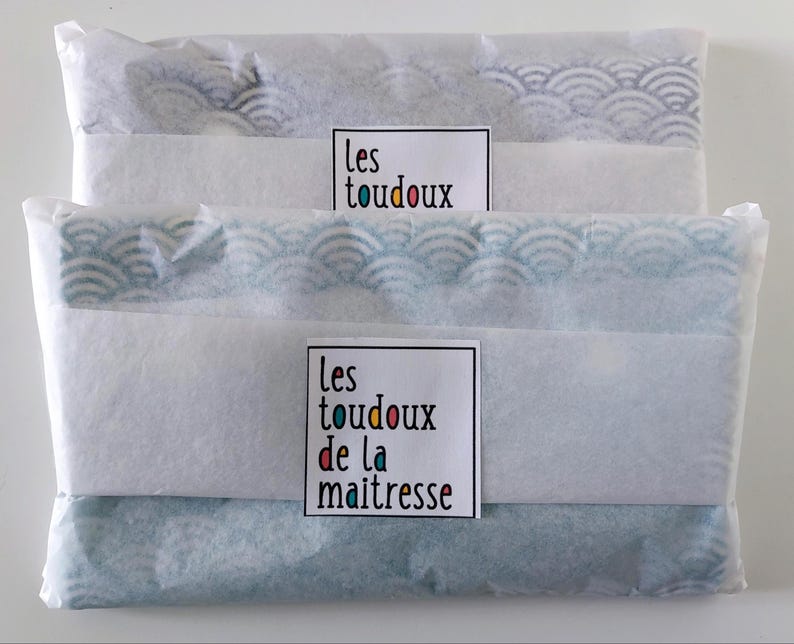 May include: Two rectangular items wrapped in white tissue paper with a blue and white wave pattern visible through the paper. Each package features a white label with the text "les toudoux" or "les toudoux de la maitresse" in black and colorful letters.