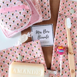 May include: A collection of beauty products, including a toothbrush, soap, and washcloths, arranged on a white surface. The items feature a pink fan-shaped pattern with black and gold accents. The box includes the text "Ma Box Beauté".