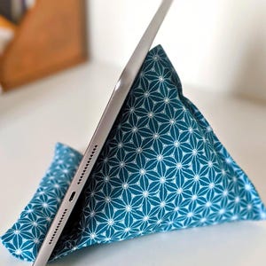 May include: A triangular fabric tablet stand featuring a teal blue background with a repeating white geometric star pattern. A silver tablet is resting against the angled side of the soft, pyramid-shaped cushion, which provides support for the device.