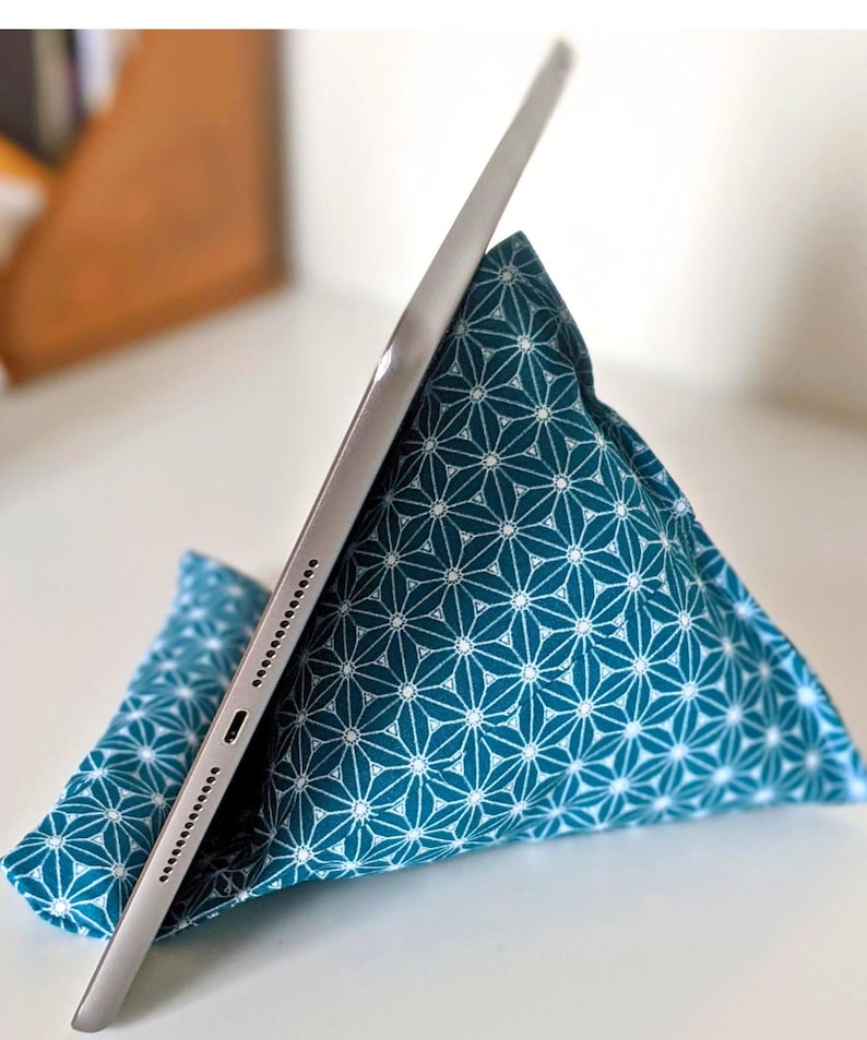 May include: A triangular fabric tablet stand featuring a teal blue background with a repeating white geometric star pattern. A silver tablet is resting against the angled side of the soft, pyramid-shaped cushion, which provides support for the device.