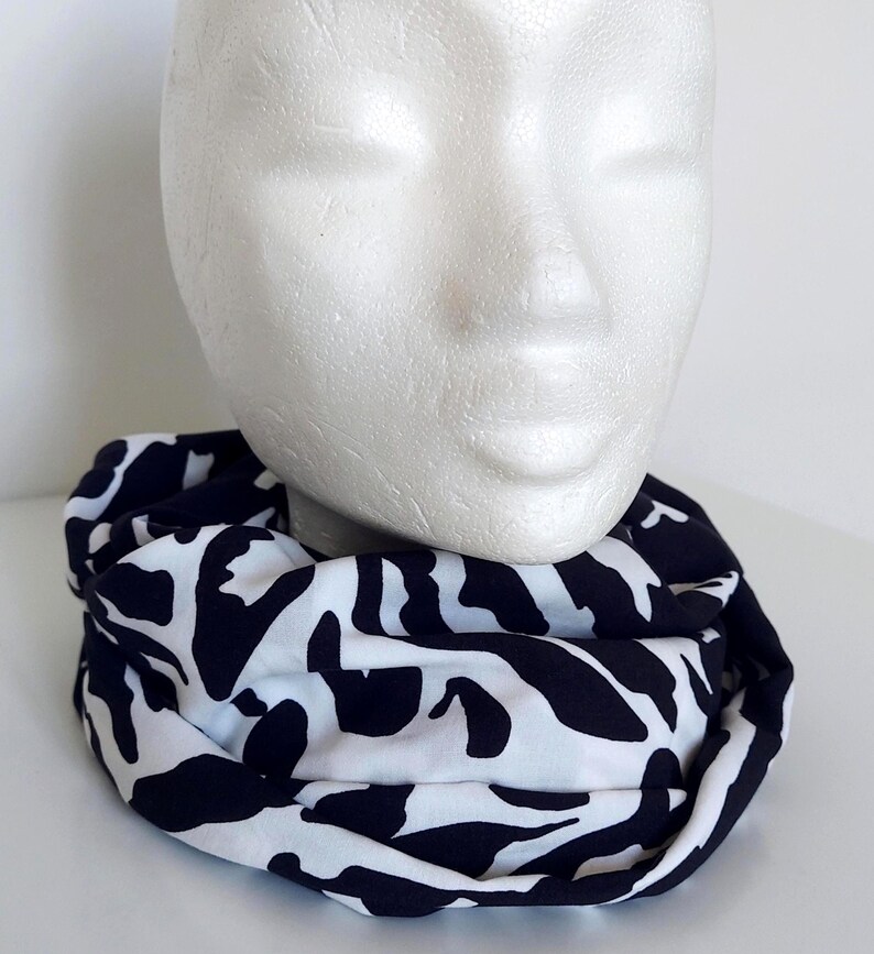 May include: An infinity scarf with a black and white abstract pattern. The scarf is draped around a white mannequin head. The design features black shapes on a white backdrop. The fabric appears to be soft and lightweight, suitable for cooler weather.