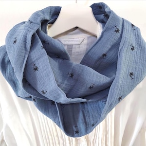 May include: A blue infinity scarf with a palm tree pattern. The scarf is draped over a white shirt on a hanger. The scarf is made of a soft, textured fabric. The palm trees are black.
