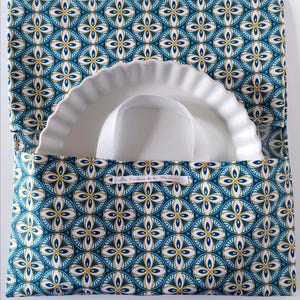 Peut inclure: Un porte-plat en tissu avec un motif géométrique circulaire bleu sarcelle, blanc et doré, conçu pour transporter un plat en céramique blanc à bords cannelés. Il possède une anse blanche et une étiquette textile Les Toiles de la Montagne.