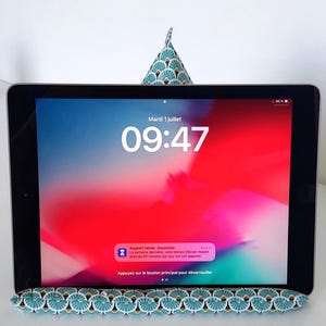 Puede incluir: Una tableta con marco negro y pantalla en tonos rojo y rosa brillantes, apoyada sobre un soporte de tela con un patrón de abanicos en color turquesa, blanco y dorado. La pantalla muestra la hora 09:47 y una notificación en francés.