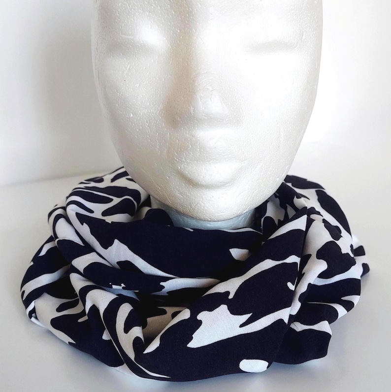 May include: A black and white patterned infinity scarf, displayed on a mannequin head. The scarf features a striking abstract design. The fabric appears soft and drapes well, suitable for various outfits and styles.