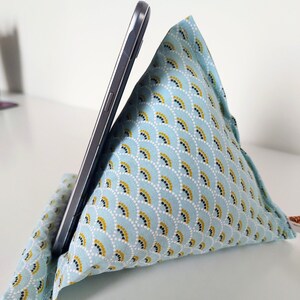 May include: A triangular fabric phone stand featuring a light blue background with a repeating pattern of white, yellow, and black semicircular arcs. A dark gray smartphone is propped against the angled side of the soft, stuffed cushion.
