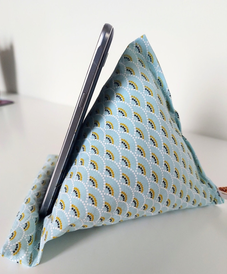 May include: A triangular fabric phone stand featuring a light blue background with a repeating pattern of white, yellow, and black semicircular arcs. A dark gray smartphone is propped against the angled side of the soft, stuffed cushion.