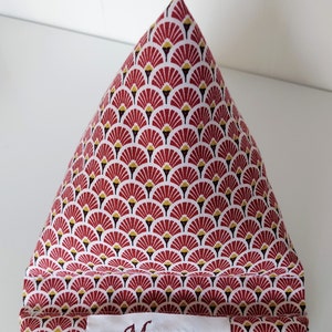 May include: A triangular-shaped fabric item with a repeating fan-like pattern in red, white, black, and gold. The fabric item has a white label with the embroidered name "Manou".