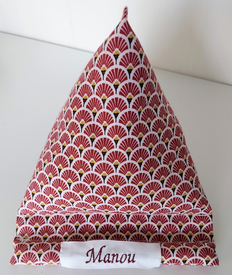 May include: A triangular-shaped fabric item with a repeating fan-like pattern in red, white, black, and gold. The fabric item has a white label with the embroidered name "Manou".