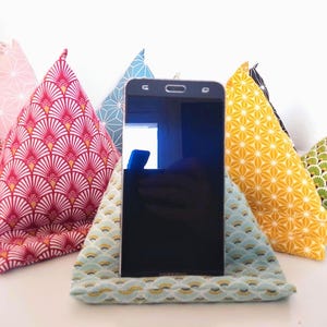 Può includere: Uno smartphone nero è appoggiato su un supporto per telefono con motivo blu chiaro, circondato da supporti in tessuto colorati. I supporti presentano vari motivi in rosso, rosa, giallo, verde e blu, creando un display vivace.