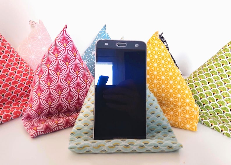 May include: A black smartphone rests in a light blue patterned fabric stand, surrounded by colorful fabric phone stands. The stands feature various patterns in red, pink, yellow, green, and blue, creating a vibrant display.