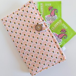 Puede incluir: Un estuche para bolsitas de té de tela con estampado de abanicos rosa y blanco, asegurado con un botón de madera. Se ven dos bolsitas de té verde con diseños de cachemira y texto. Las bolsitas de té dicen "A BAG OF OUR SENSATIONAL THE VERT CHA".