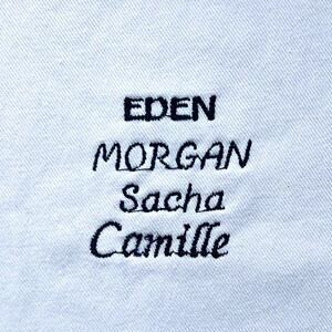 May include: Close-up of a white fabric with black embroidered names: EDEN, MORGAN, Sacha, and Camille. The names are in a mix of uppercase and lowercase letters, stitched in a cursive style. The fabric has a subtle, textured appearance.