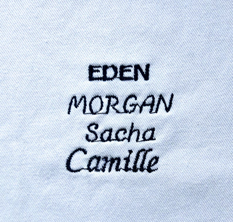May include: Close-up of a white fabric with black embroidered names: EDEN, MORGAN, Sacha, and Camille. The names are in a mix of uppercase and lowercase letters, stitched in a cursive style. The fabric has a subtle, textured appearance.