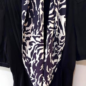 May include: A black dress with short sleeves and a navy and white patterned scarf. The scarf features an abstract design. The dress is hanging on a black hanger.