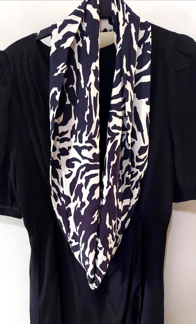 May include: A black dress with short sleeves and a navy and white patterned scarf. The scarf features an abstract design. The dress is hanging on a black hanger.