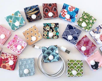 Puede incluir: Una colección de organizadores de cables cuadrados de tela con varios patrones coloridos, cada uno asegurado con un botón de presión de plástico blanco. Un organizador está envuelto alrededor de un cable de carga blanco, rodeado por los demás.