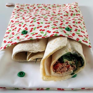 Puede incluir: Una envoltura de sándwich de tela blanca con un estampado de cerezas rojas, que contiene un wrap relleno de verduras y carne. La envoltura se asegura con cierres de presión verdes. El wrap está abierto, revelando el contenido.