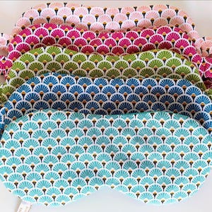 May include: A stack of eye masks in various colors, including pink, fuchsia, green, blue, and teal. Each mask features a repeating fan-shaped pattern in white and gold. The masks are made of fabric and have elastic straps.