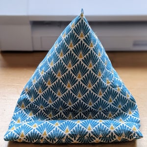 May include: A triangular fabric stand featuring a repeating pattern of teal fan-shaped motifs with small gold triangles on a white background. The object has a wide base and a pointed top, designed with a horizontal ledge at the front for support.