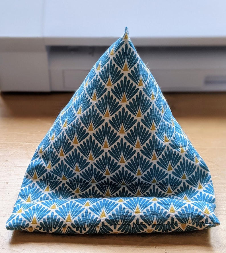 May include: A triangular fabric stand featuring a repeating pattern of teal fan-shaped motifs with small gold triangles on a white background. The object has a wide base and a pointed top, designed with a horizontal ledge at the front for support.
