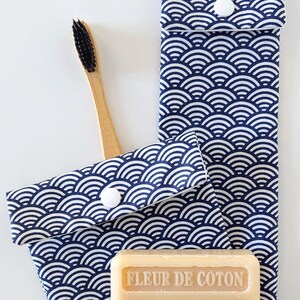 May include: A bamboo toothbrush with black bristles, a bar of soap, and two navy blue and white wave-patterned fabric pouches. The soap is labeled "FLEUR DE COTON" and "FABRIQUÉ A MARSEILLE". The pouches have white button closures.