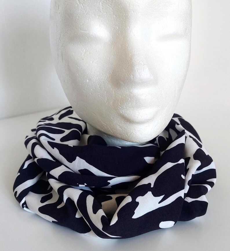 May include: A navy and white patterned infinity scarf, displayed on a mannequin head. The scarf's design features irregular white shapes against a dark blue backdrop. The fabric appears soft and drapes well, suggesting a comfortable accessory.