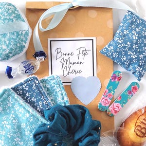 May include: A gift set with a brown paper bag tied with a pale blue ribbon. The set includes floral print items, a scrunchie, nail files with rose designs, a heart-shaped card, and a wrapped sweet. The card reads "Bonne Fête Maman Chérie."