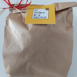 May include: A brown paper gift bag featuring a yellow square tag with the text "les toudoux de la maitresse" and a small gold star sticker. The bag is tied at the top with a red and natural-colored raffia ribbon bow, creating a simple presentation.