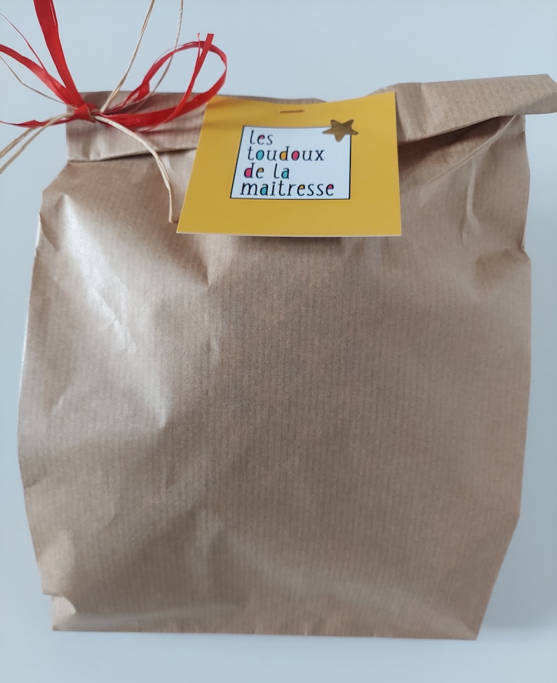 May include: A brown paper gift bag featuring a yellow square tag with the text "les toudoux de la maitresse" and a small gold star sticker. The bag is tied at the top with a red and natural-colored raffia ribbon bow, creating a simple presentation.