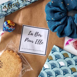 May include: A gift box featuring a teal scrunchie, patterned fabric, a cookie, and a lollipop. The box includes a teal ribbon with gold text and a card that reads "La Box Pour Elle".