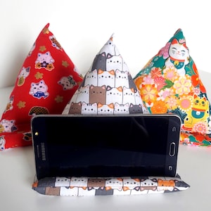 May include: Three triangular fabric phone stands featuring cat-themed patterns. One is red with white cats, one is white with a grid of cat faces, and one is green with floral motifs and beckoning cats. A black smartphone rests on the white stand.