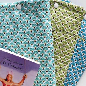 Può includere: Un gruppo di copertine per libri in tessuto verde acqua, verde e blu, ciascuna con un motivo a forma di ventaglio. Le copertine sono fissate con bottoni bianchi. Un libro con il titolo "Je L'aimais" è parzialmente visibile.