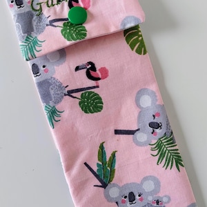 May include: A rectangular fabric pouch in light pink featuring a repeating pattern of grey koalas, green tropical leaves, and small toucans. The flap is secured with a green plastic snap button and displays the embroidered name Gabriela.