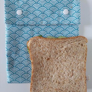 May include: A rectangular fabric sandwich bag featuring a teal and white wave pattern. The bag has a fold-over flap secured by two white plastic snap buttons. A single slice of whole wheat bread is positioned in front of the bag.