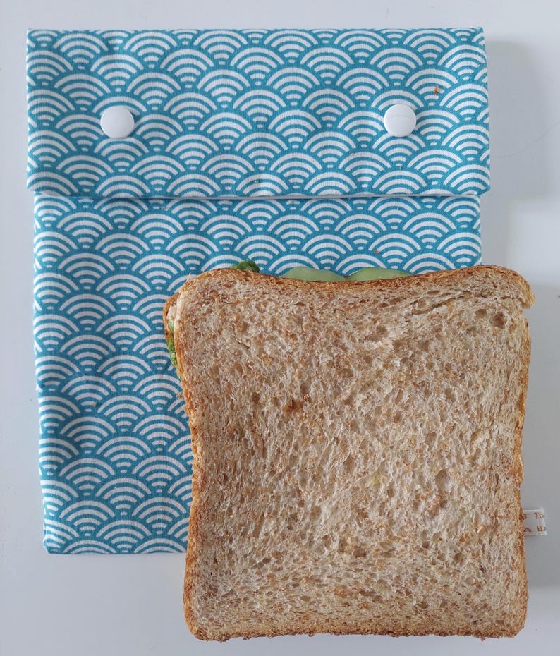 May include: A rectangular fabric sandwich bag featuring a teal and white wave pattern. The bag has a fold-over flap secured by two white plastic snap buttons. A single slice of whole wheat bread is positioned in front of the bag.