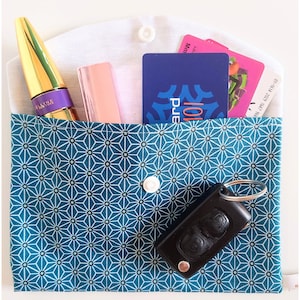 May include: A blue fabric pouch with a white geometric pattern. The pouch has a white zipper and a white button. The pouch is filled with various items, including a gold tube of mascara, a pink lipstick, a blue and white card, a pink card, and a black car key with a silver key ring.