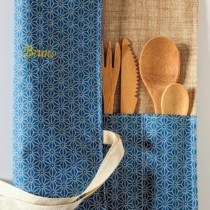 Op de afbeelding: Een blauw-wit geometrisch patroon bestekrol met houten keukengerei. De rol is open en toont een vork, mes en twee lepels. Het woord "Bruno" is in het geel geborduurd.