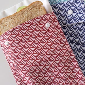 May include: A red and white fabric sandwich bag featuring a repeating arched wave pattern and a white plastic snap closure. A whole wheat sandwich is partially visible inside the bag. A second blue and white bag is partially visible behind it.