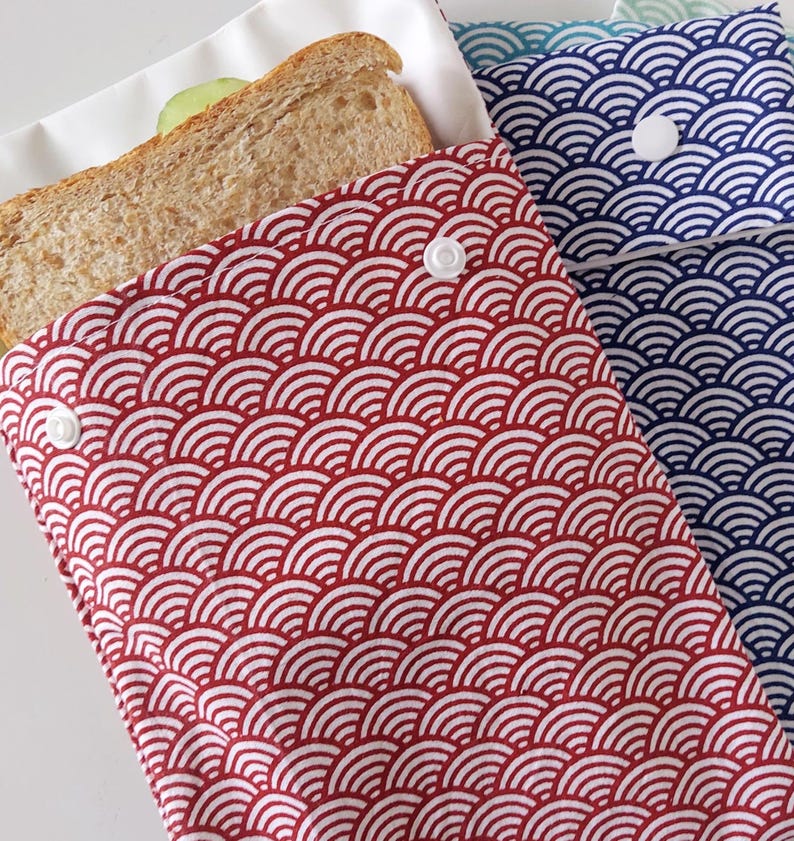 May include: A red and white fabric sandwich bag featuring a repeating arched wave pattern and a white plastic snap closure. A whole wheat sandwich is partially visible inside the bag. A second blue and white bag is partially visible behind it.